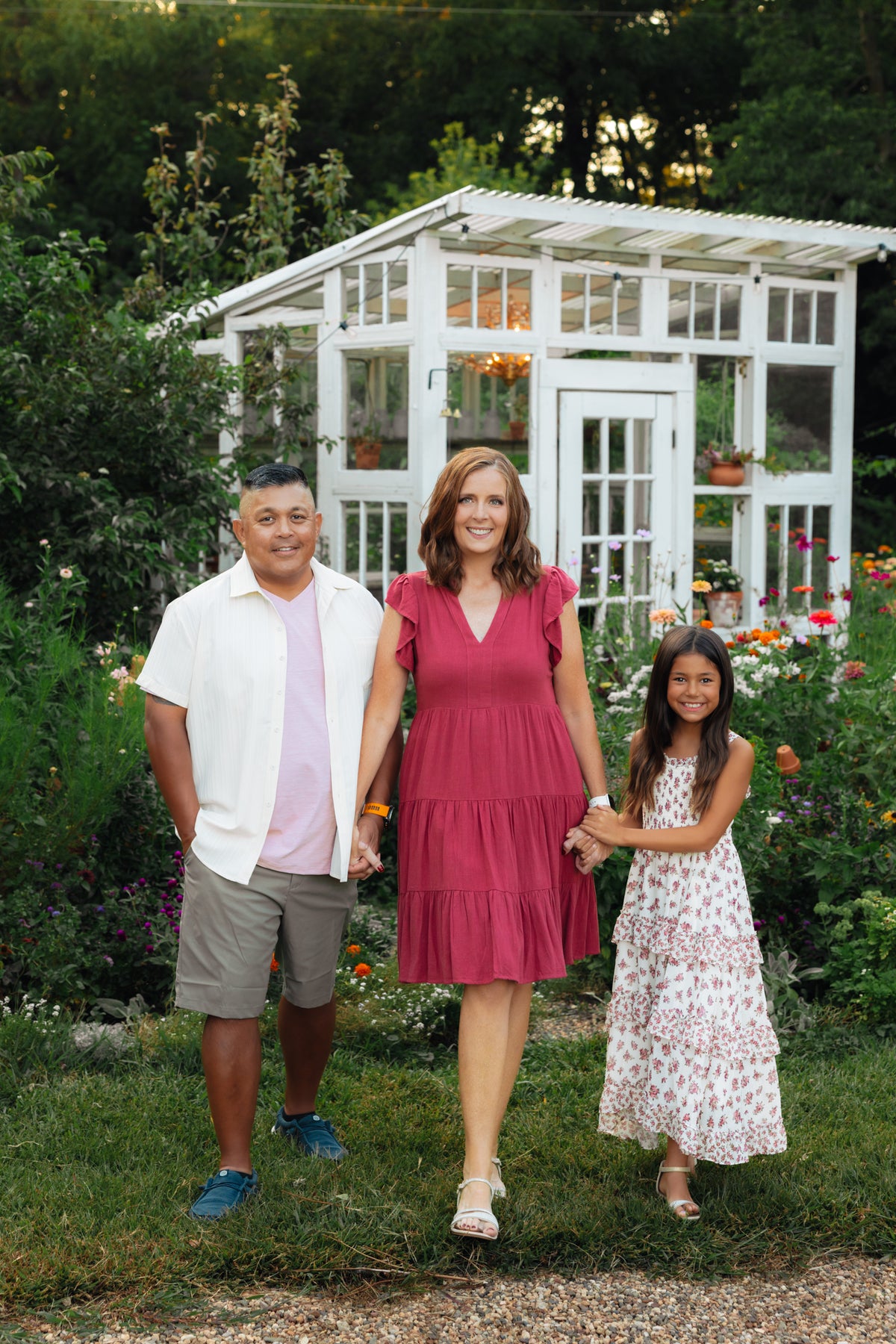 the Juli Camarin family stands in a garden in front of a white greenhouse. The mother wears a sleeveless dusty rose pink tiered dress, the father wears a white short-sleeve button-up shirt over a light pink t-shirt and khaki shorts, and the daughter wears a white floral print maxi dress.