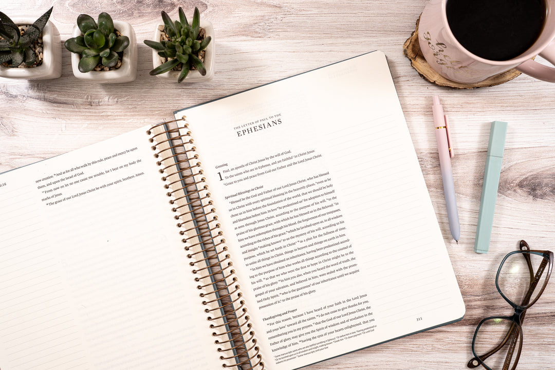 Open notebook on a wooden desk with a coffee cup, glasses, and plants.
