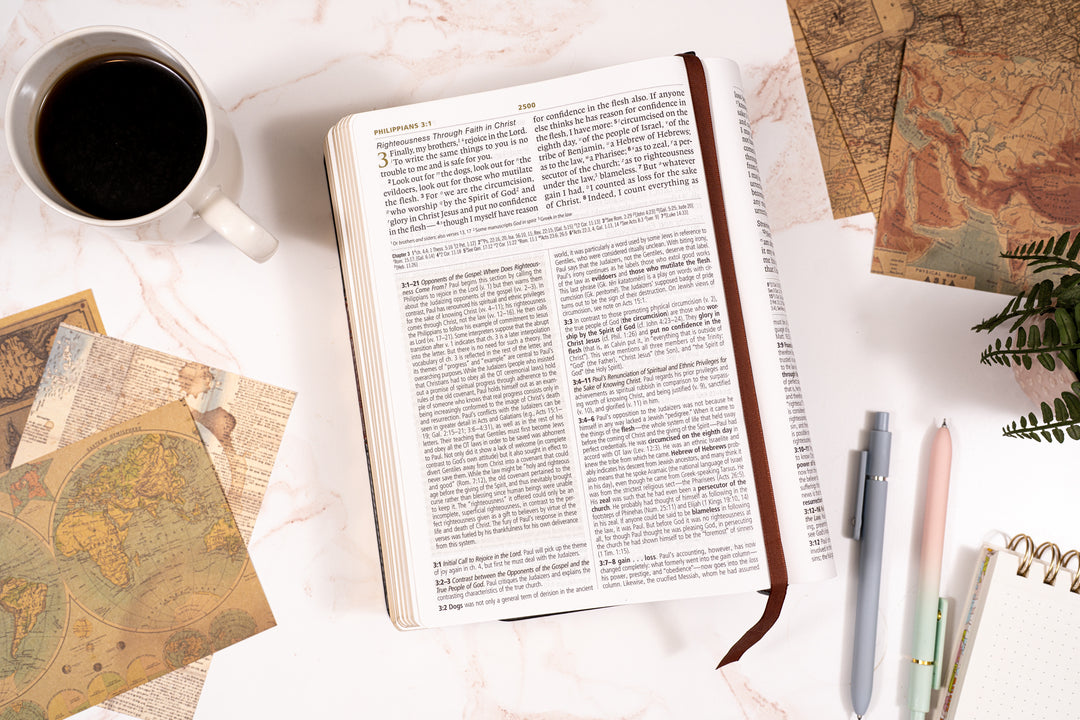 Open bible on a table with a cup of coffee, maps, and pens.