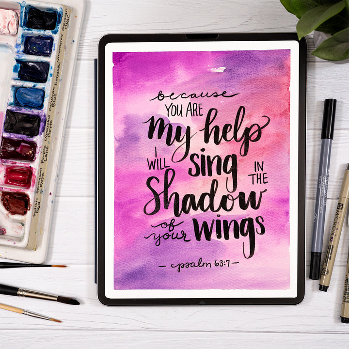 Tablet with motivational quote on a watercolor background, surrounded by art supplies on a white surface.